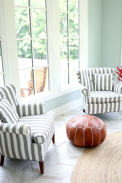Living room with striped armchairs and a leather pouf in front of large windows.
