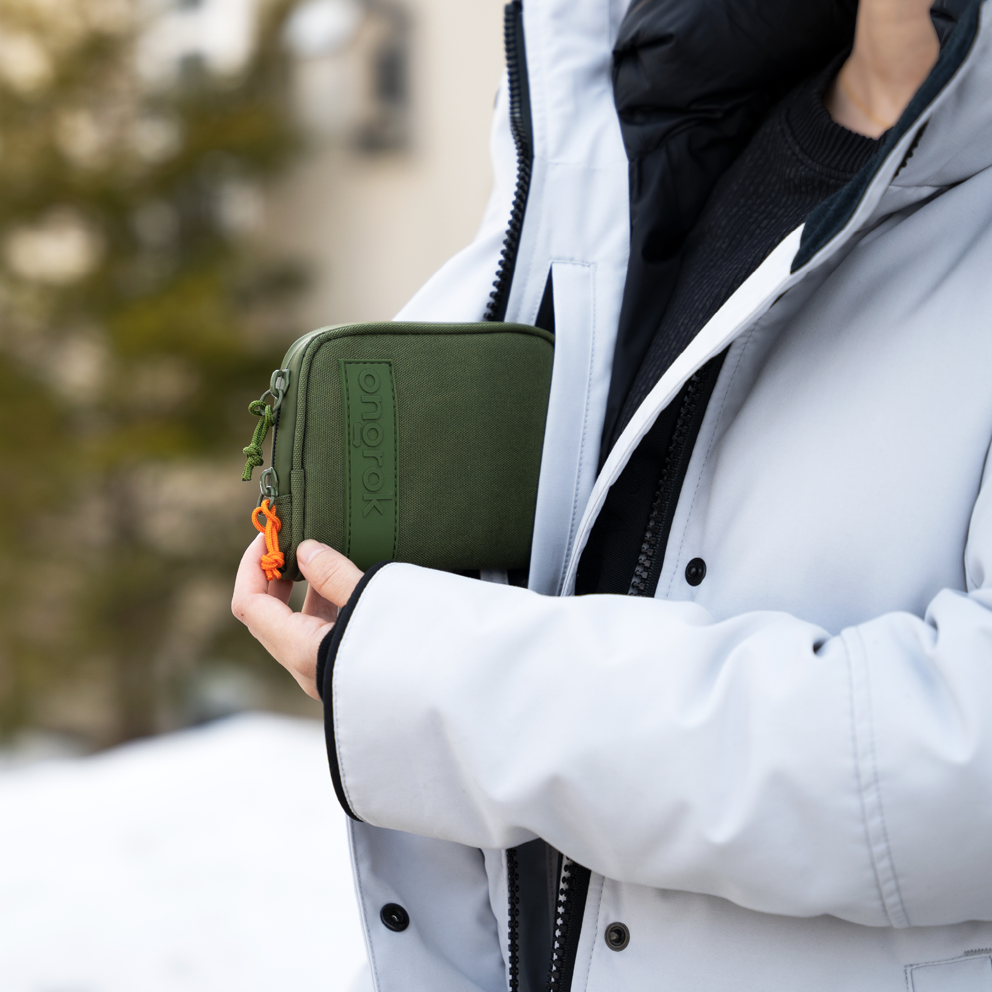 Person holding a green pouch with a brand logo, wearing a white jacket outdoors.