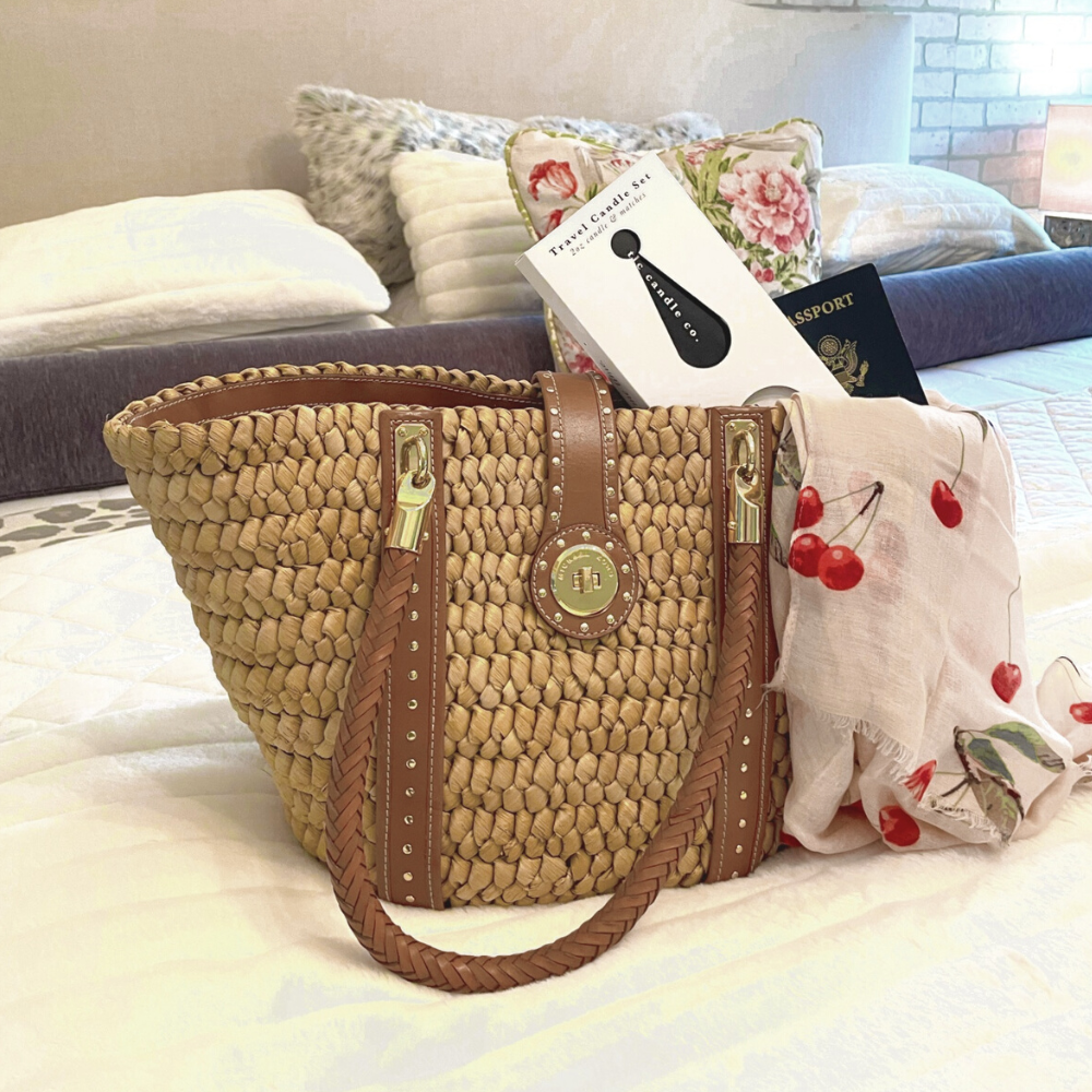 Woven handbag with brown handles on a bed with pillows and a book.