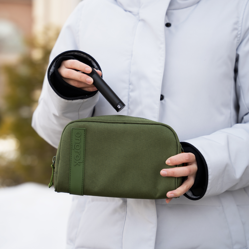 Person holding a green toiletry bag with a black item, wearing a white coat outdoors.