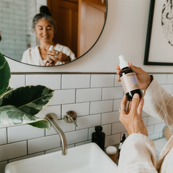 Person holding a bottle of skincare product in front of a mirror in a bathroom.