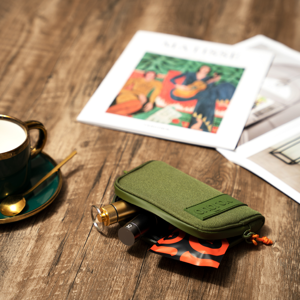 Green pouch with vape device on a wooden table with a cup and saucer and magazine in the background