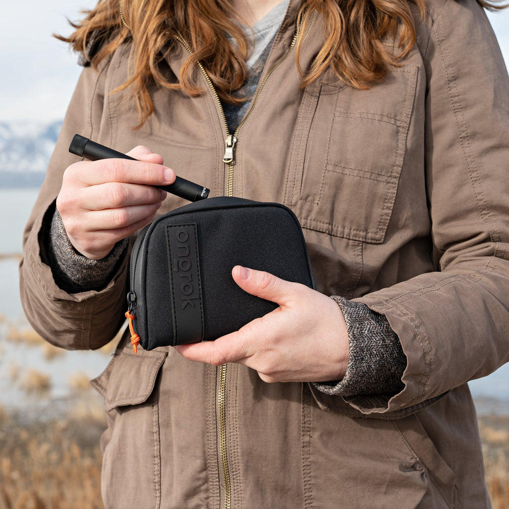Person holding a black carrying case with a pen, wearing a brown jacket outdoors.