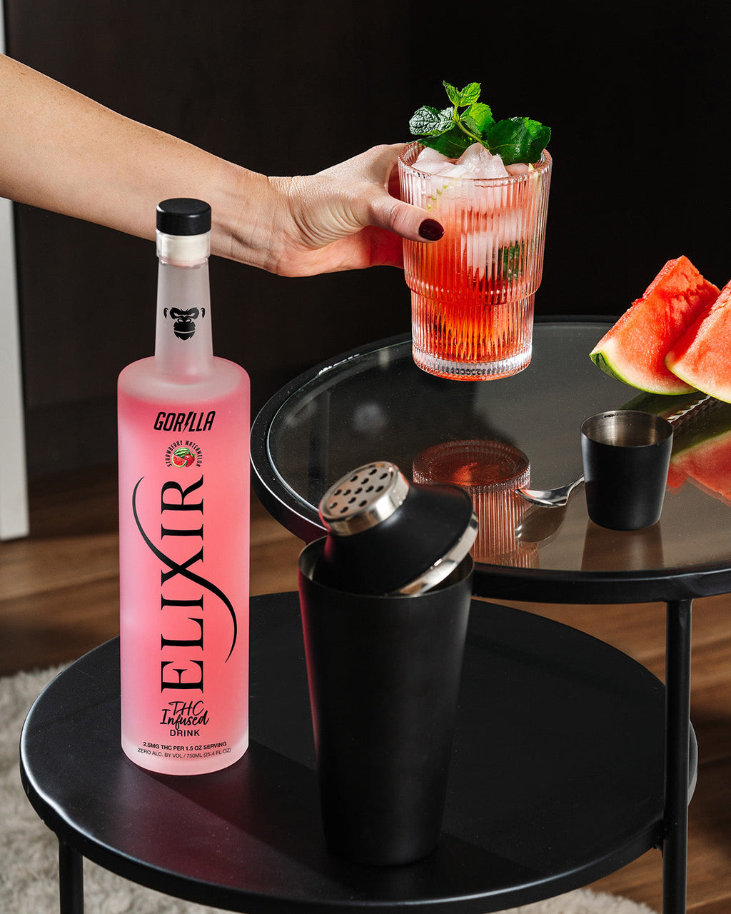 Person holding a cocktail with Elixir Gorilla bottle and sliced watermelon on a table.