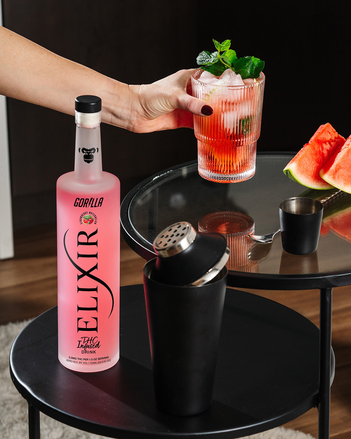 Person holding a cocktail with Elixir Gorilla bottle and sliced watermelon on a table.