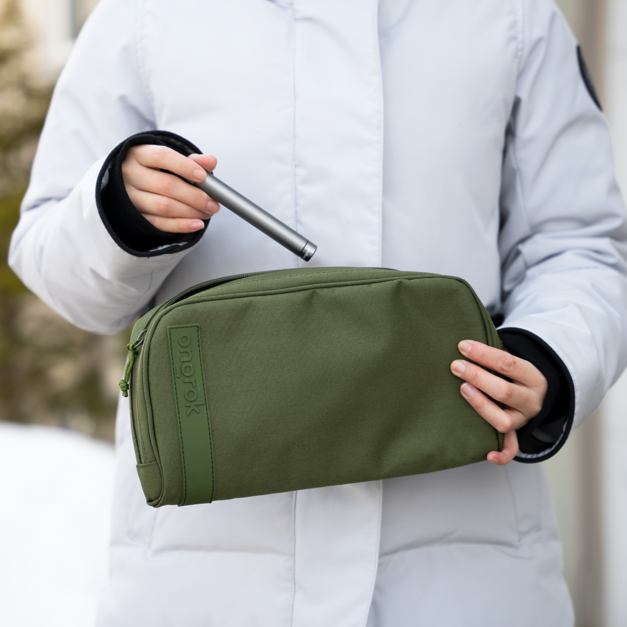 Person holding a green toiletry bag with a pen, wearing a light gray coat.