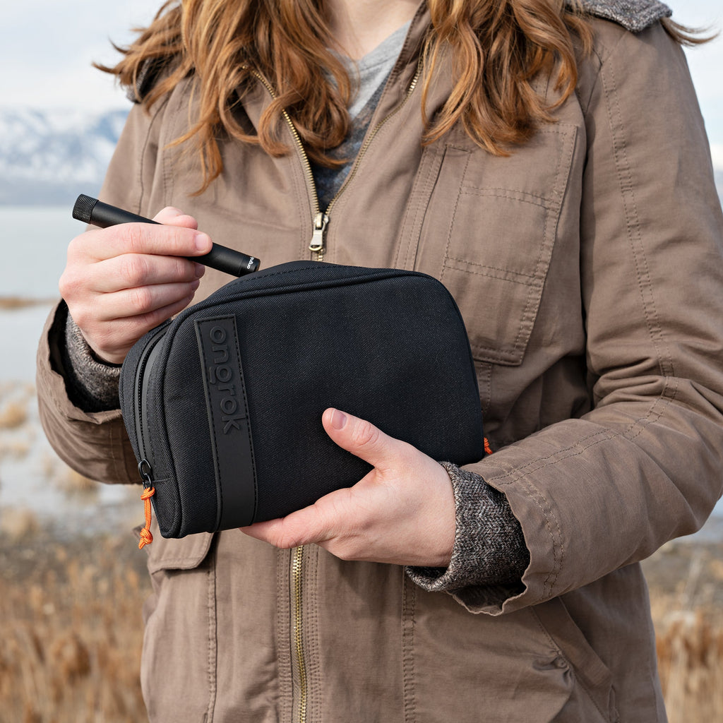 Person holding a black toiletry bag with a pen, outdoors.