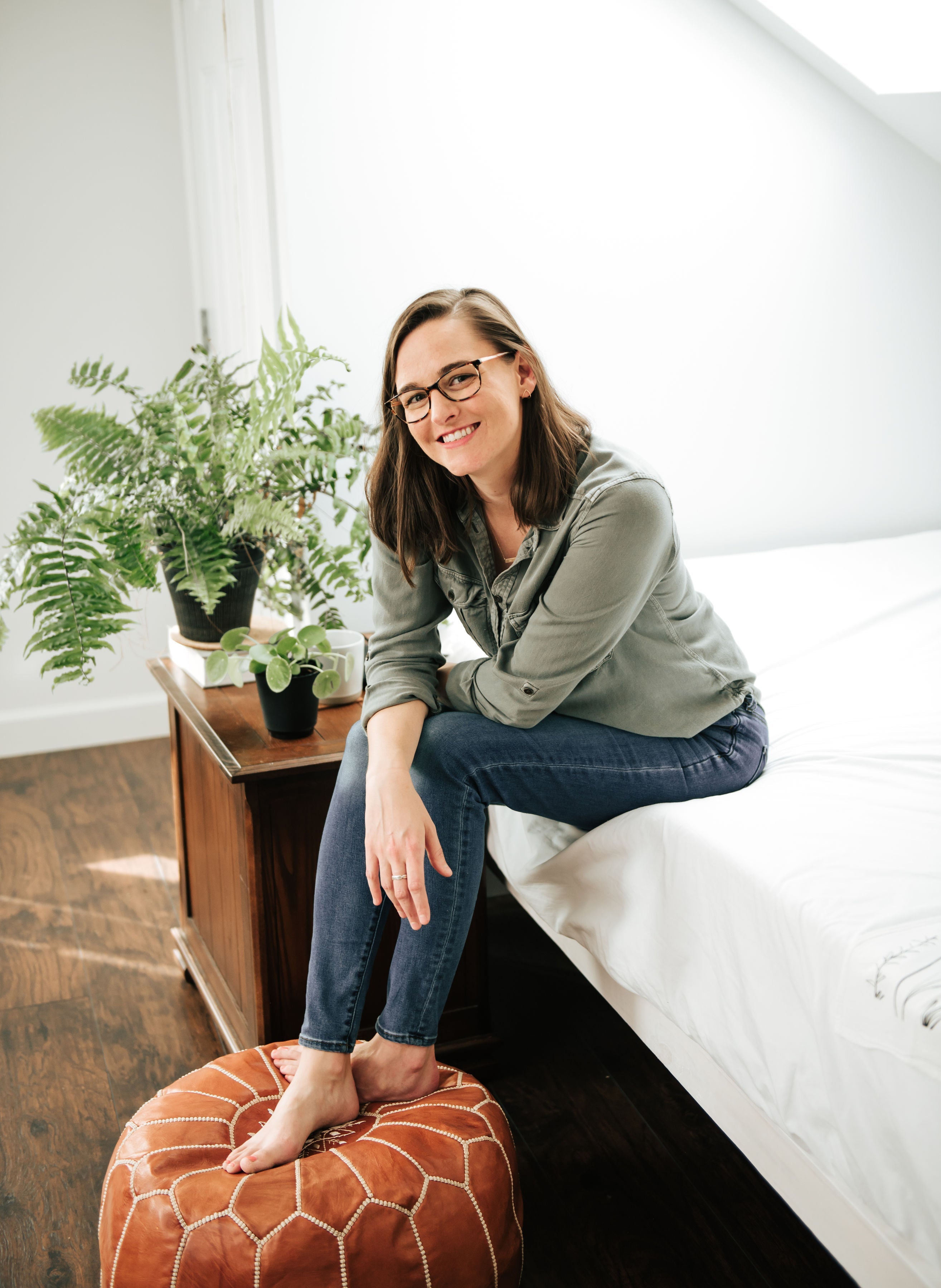 Woman sitting on a white bed in a bedroom with plants and a brown ottoman.
