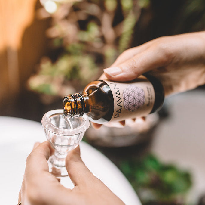 Person pouring a liquid from a bottle labeled 'PAAVAN' into a small glass container with a blurred natural background.