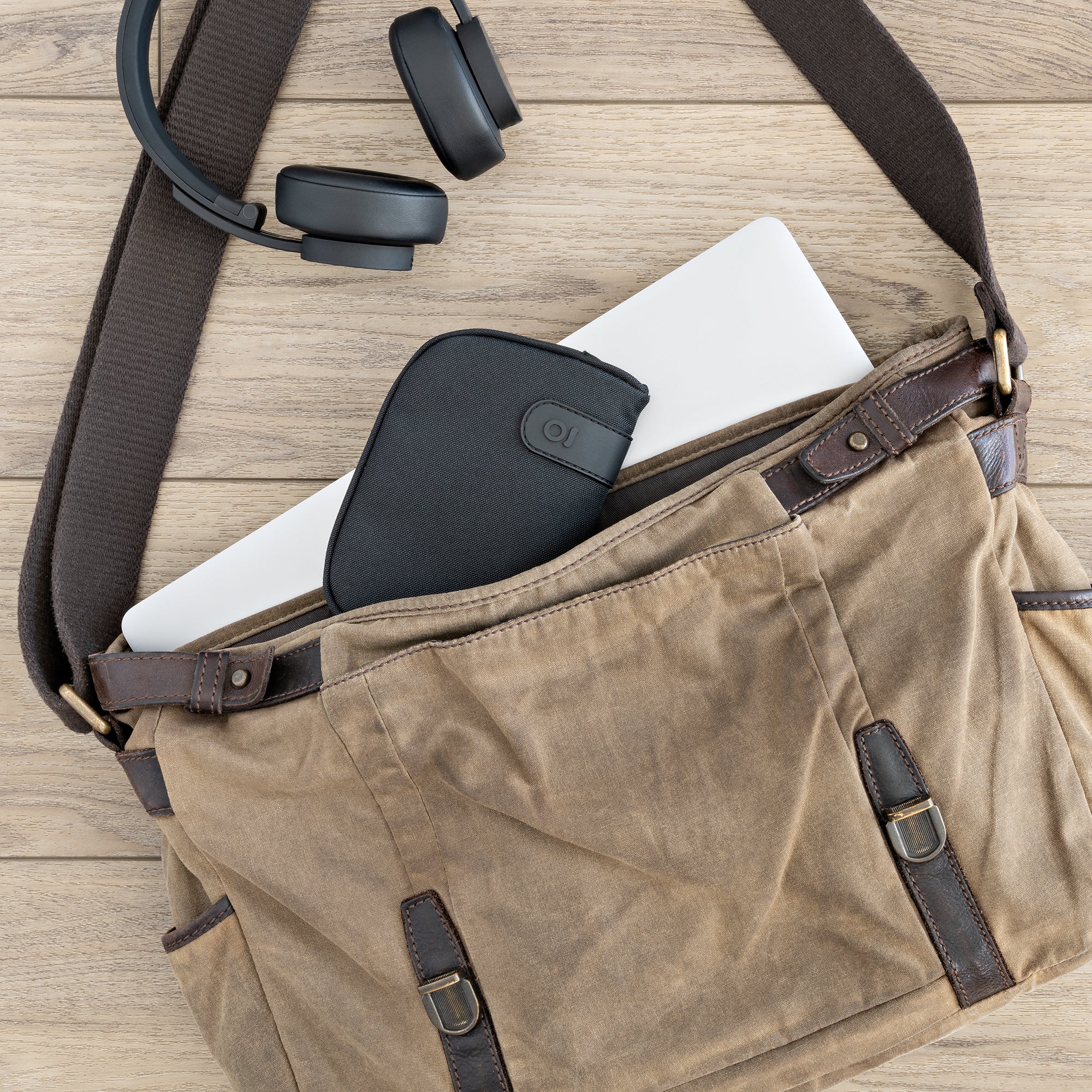 Brown leather bag with a black strap, containing a laptop and black case on a wooden surface.